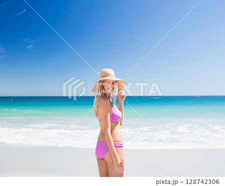 Young woman standing in profile on sandy beach, with pink bikini and wide?brimmed straw sun hat Young woman standing in profile on sandy beach, with pink bikini and wide?brimmed straw sun hat 128742306