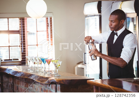 African American bartender shaking cocktail shaker behind wood bar with martini glasses, copy space 128742356