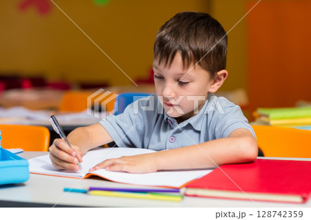 Boy writing at school desk in elementary classroom, with pen open notebook red folder pencil case 128742359