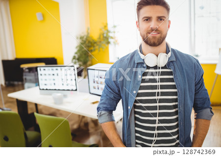 Man in mid 20s standing in modern office, with headphones, monitors and coffee mug, copy space Man in mid 20s standing in modern office, with headphones, monitors and coffee mug, copy space 128742369