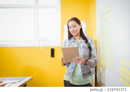 Asian woman standing near center holding tablet in bright creative office, with sticky notes Asian woman standing near center holding tablet in bright creative office, with sticky notes 128742371