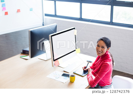 Young Chinese woman creating art on drawing tablet in office, with dual monitors and sticky notes Young Chinese woman creating art on drawing tablet in office, with dual monitors and sticky notes 128742376