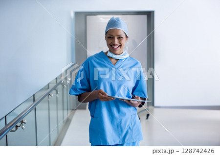 bi-racial young nurse standing in bright hospital corridor, holding tablet and smiling 128742426