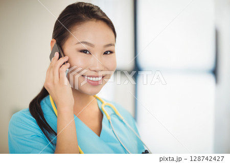 Asian female nurse talking on smartphone in clinic, with yellow stethoscope and bright window 128742427