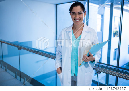 Female doctor leaning on railing in hospital corridor, holding blue folder, smiling, copy space 128742437
