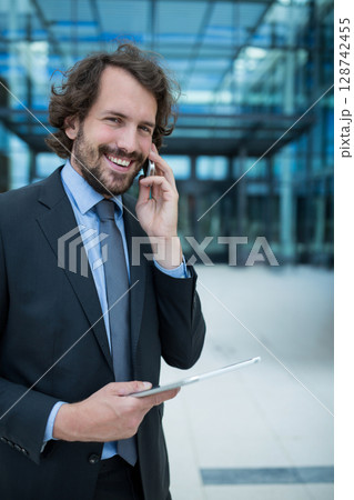 Businessman talking on smartphone while holding tablet outside modern office plaza, copy space Businessman talking on smartphone while holding tablet outside modern office plaza, copy space 128742455