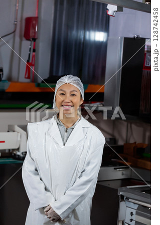 Asian woman standing and smiling inside food processing facility, with lab coat hairnet gloves 128742458