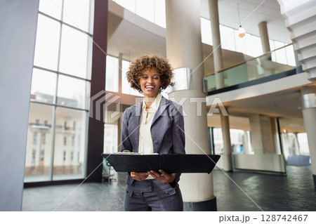 African American businesswoman standing in office atrium with black portfolio folder, smiling African American businesswoman standing in office atrium with black portfolio folder, smiling 128742467
