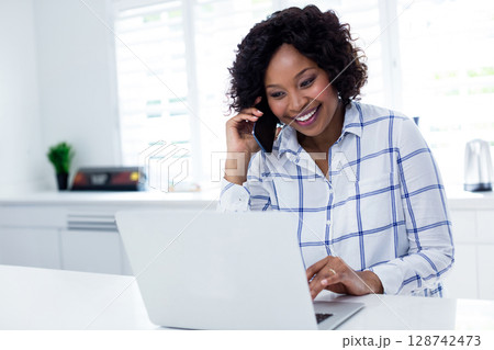 African American woman holding smartphone and typing on laptop in bright kitchen African American woman holding smartphone and typing on laptop in bright kitchen 128742473