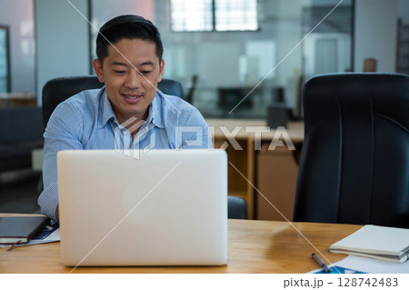 Asian man sitting at wooden desk in open-plan office, working on silver laptop with white notepad 128742483