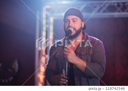 Man gripping mic stand and singing on concert stage, with truss lights and blurred guitar headstock 128742546