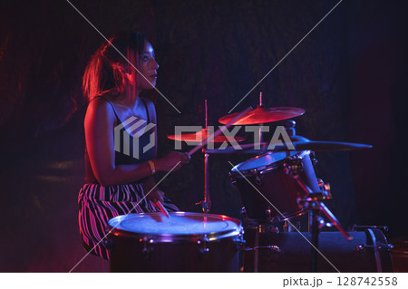 Young adult Asian female drummer performing on stage with drum kit under red and blue lights 128742558