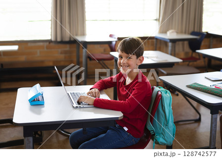 Young boy typing on silver laptop in school classroom, with blue pencil case and teal backpack 128742577