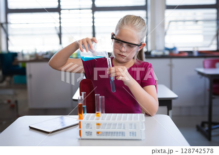 Girl pouring blue liquid into test tube at lab desk, with test tube rack and tablet 128742578