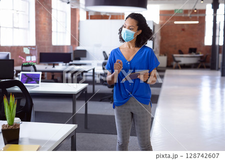 Woman wearing surgical mask using tablet and stylus in open-plan office with potted succulent 128742607