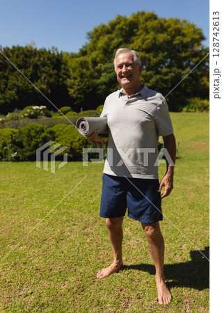 Senior man standing barefoot in backyard garden, holding gray yoga mat under left arm Senior man standing barefoot in backyard garden, holding gray yoga mat under left arm 128742613