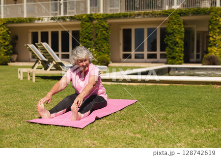 Senior woman stretching forward on pink yoga mat in backyard garden, with pool and lounge chairs 128742619