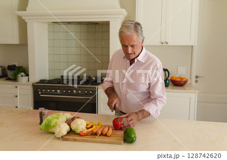 Senior man slicing red bell pepper on wooden cutting board in modern kitchen, with fresh vegetables Senior man slicing red bell pepper on wooden cutting board in modern kitchen, with fresh vegetables 128742620