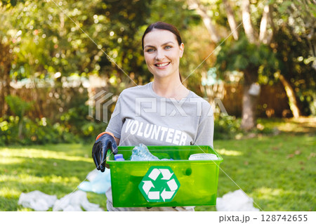 Woman volunteer holding green recycling bin in sunny park, smiling with plastic bottles and bags 128742655