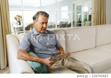 Senior man sitting on sofa in bright living room, holding tablet with turquoise pillow, copy space 128742687