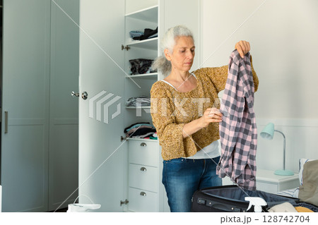 Senior woman sorting clothes from black suitcase in minimalist bedroom, featuring built-in wardrobe Senior woman sorting clothes from black suitcase in minimalist bedroom, featuring built-in wardrobe 128742704