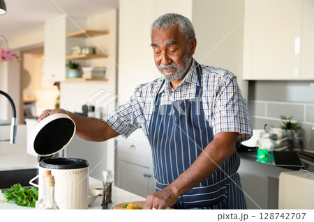 Senior man lifting compost bin lid while chopping bell peppers and herbs at modern kitchen island Senior man lifting compost bin lid while chopping bell peppers and herbs at modern kitchen island 128742707