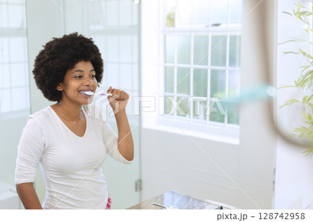 African American woman brushing teeth at bathroom sink with toothbrush and plant, copy space 128742958