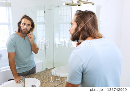 Young man touching beard and examining reflection at bathroom mirror, with sink, copy space 128742960