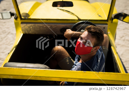 Male wearing red face mask driving yellow dune buggy on sandy terrain, with steering wheel Male wearing red face mask driving yellow dune buggy on sandy terrain, with steering wheel 128742967