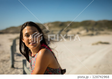 Woman leaning on wooden fence at sandy coastal dune, smiling at camera with breeze, copy space 128742968