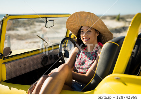 Young adult woman wearing straw hat sitting in dune buggy on coastal dunes, with calm joy 128742969