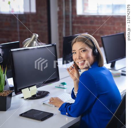 Asian woman typing on keyboard and talking on headset at office desk with monitors, copy space Asian woman typing on keyboard and talking on headset at office desk with monitors, copy space 128742976