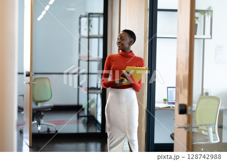 Young African American woman standing in office corridor, holding yellow folder beside open laptop Young African American woman standing in office corridor, holding yellow folder beside open laptop 128742988