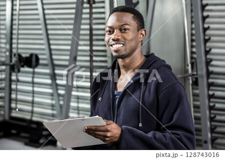 Young African American gym trainer standing in gym, with clipboard, weight machines and squat rack 128743016