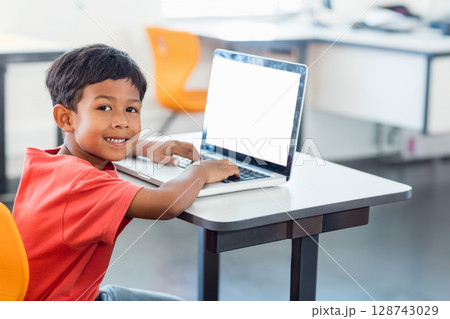 Boy typing on laptop at desk smiling at camera in classroom, with orange chairs, copy space 128743029