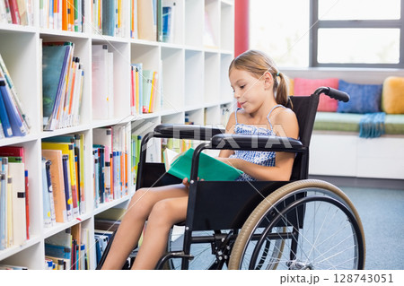 African American teenage girl sitting in manual wheelchair in library aisle reading open green book African American teenage girl sitting in manual wheelchair in library aisle reading open green book 128743051