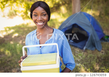 Young African American woman holding cooler near camping tent at campsite, enjoying sunny day 128743059