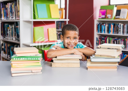Boy leaning on three stacks of books in school library, with gray table and colorful binders 128743083