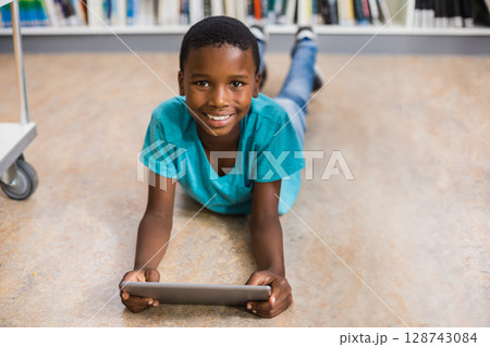 African American boy lying on floor in library with white bookshelf, holding tablet near book cart 128743084