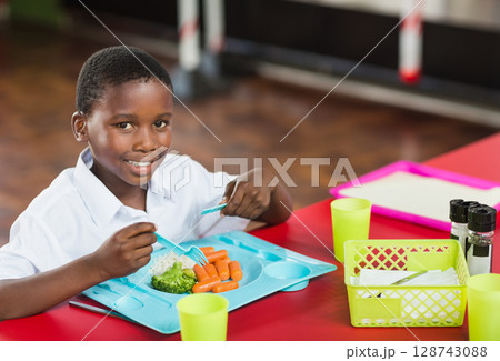 African American boy eating lunch at red cafeteria table, using blue tray with broccoli and carrots African American boy eating lunch at red cafeteria table, using blue tray with broccoli and carrots 128743088