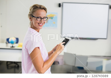 Female teacher standing in bright classroom holding tablet, with globe and world map, copy space 128743089