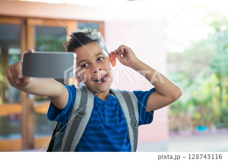 African American boy taking smartphone selfie at school entrance wearing backpack and playful face 128743116