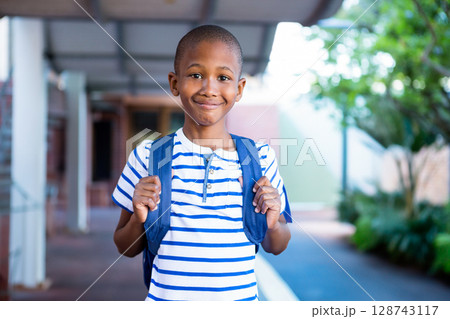 African American boy standing under covered walkway at school, holding blue backpack and smiling African American boy standing under covered walkway at school, holding blue backpack and smiling 128743117