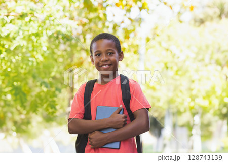 Diverse male child holding blue tablet, wearing black backpack in sunlit park, smiling at camera Diverse male child holding blue tablet, wearing black backpack in sunlit park, smiling at camera 128743139