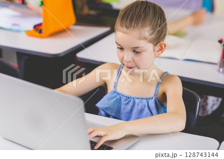 Female child working on silver laptop at school desk in classroom, with orange pencil case 128743144