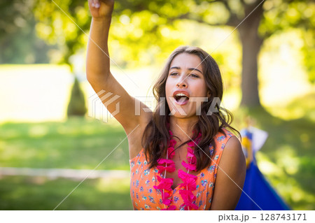 Woman raising arm and cheering in sunlit park, with pink flower lei at outdoor gathering Woman raising arm and cheering in sunlit park, with pink flower lei at outdoor gathering 128743171
