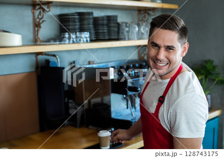 Asian male barista standing behind café counter, holding cup near espresso machine, copy space Asian male barista standing behind café counter, holding cup near espresso machine, copy space 128743175