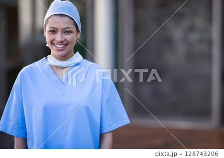 Female nurse standing by hospital entrance wearing light blue scrubs and cap with mask around neck 128743206