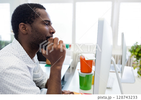 African American man sitting at office desk, gazing at monitor with storage bins, plant, copy space African American man sitting at office desk, gazing at monitor with storage bins, plant, copy space 128743233