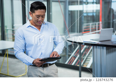 Asian man standing examining tablet in office space with metal staircase and laptop, copy space 128743276